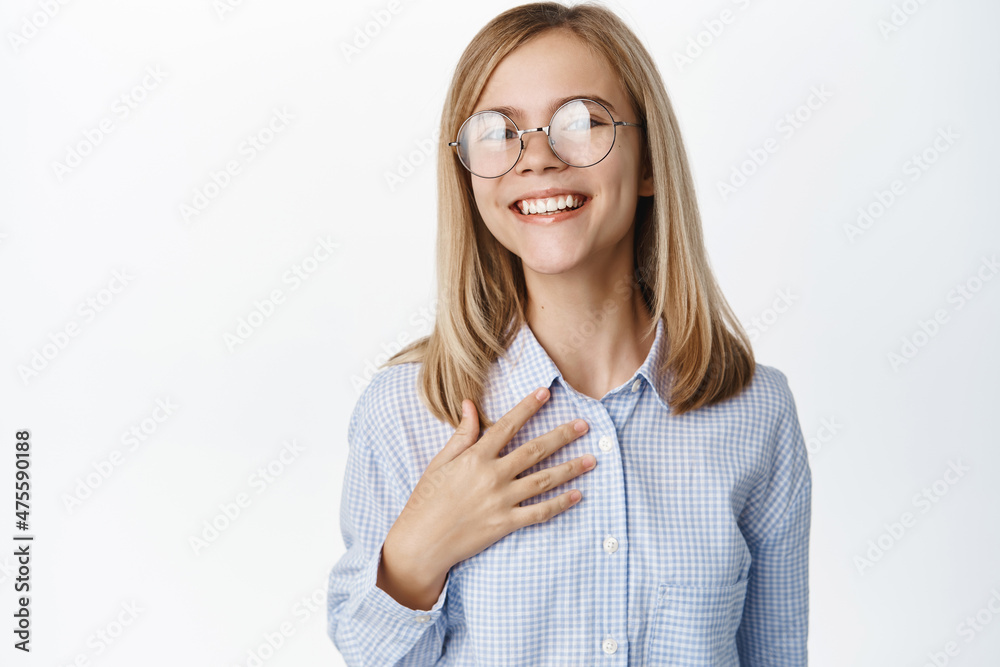 Portrait of smiling cute blond girl, child in glasses and shrit, holding hand on chest with happy face expression, standing over white background Portrait of smiling cute blond girl, child in glasses and shrit, holding hand on chest with happy face expression, standing over white background