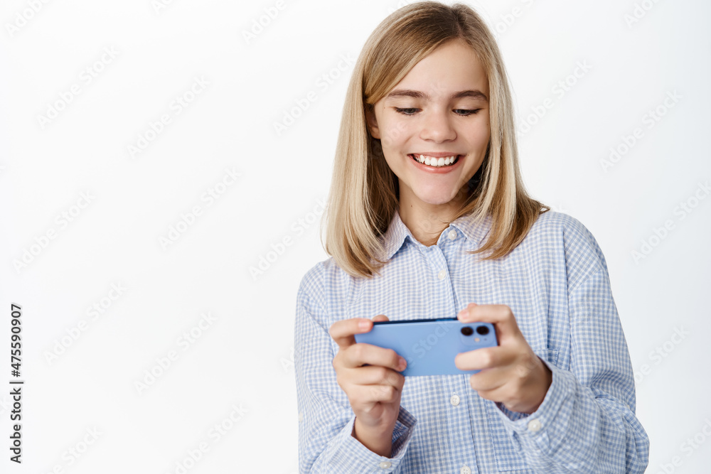 Happy cute little girl, child playing mobile video games and smiling, laughing, standing in casual clothes over white background Happy cute little girl, child playing mobile video games and smiling, laughing, standing in casual clothes over white background