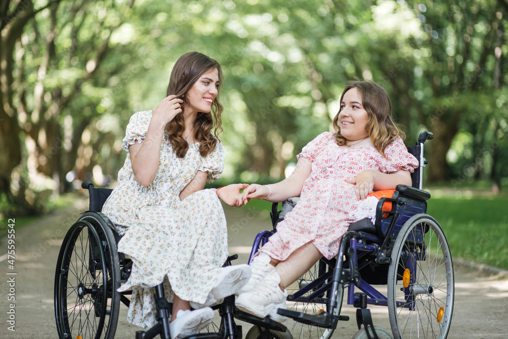 Female friends with chronic health condition spending free time together at green summer park. Charming women in wheelchairs holding hands and smiling to each other.