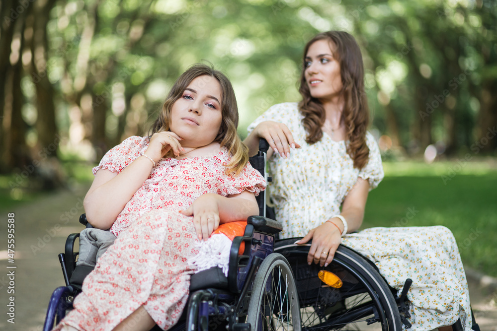 Pleasant women with disability in sitting in wheelchairs close to each other and looking at the camera. Female friends in summer dresses enjoying leisure time together at green park. Pleasant women with disability in sitting in wheelchairs close to each other and looking at the camera. Female friends in summer dresses enjoying leisure time together at green park.