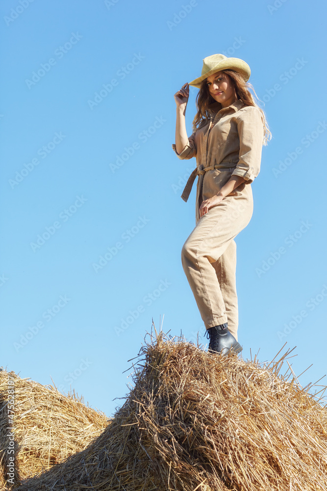Young blonde woman in a beige jumpsuit stands on on the rolls of hay. Young blonde woman in a beige jumpsuit stands on on the rolls of hay.
