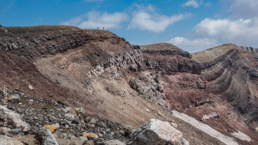 A fragment of the slope of the volcano crater. The layered texture of the soil is visible, stones of different sizes. On the edge, against the blue sky, - tiny silhouettes of tourists. Kamchatka