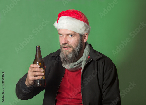 a man with a beard in a red sweater, a Harrington jacket and a Santa Claus hat stands with a bottle of beer in his hand looks questioningly with a cunning and stern look photo