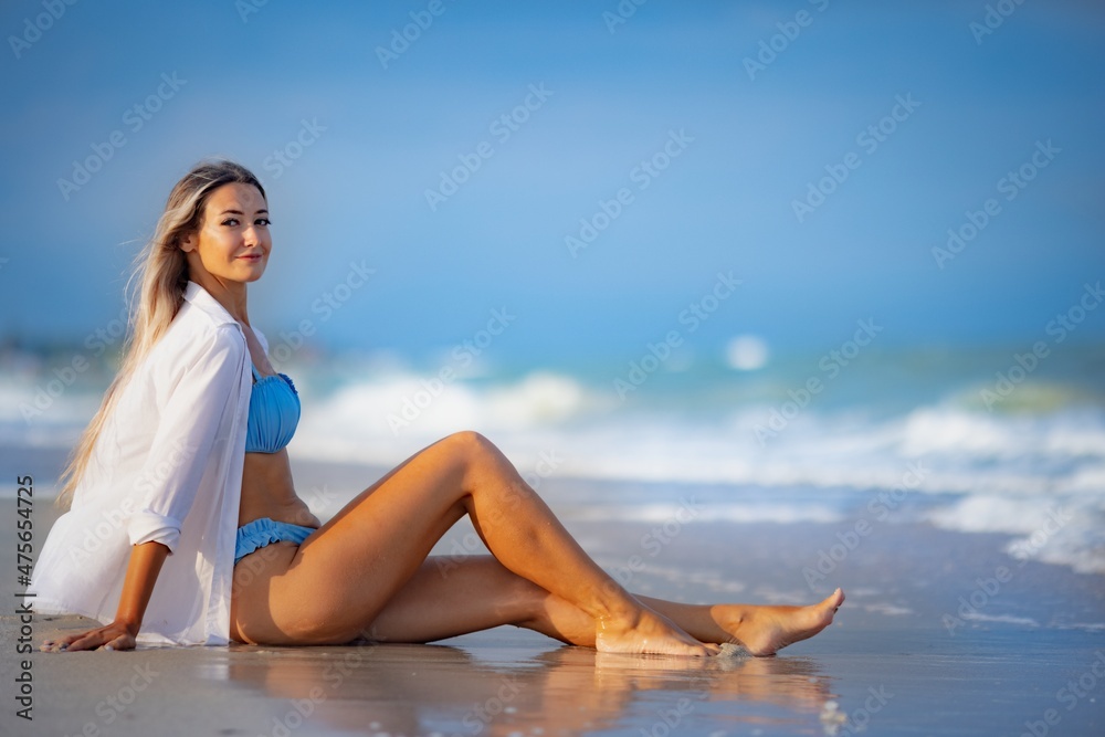 A girl in a bluish swimsuit and shirt sits near the water edge and peers into the horizon