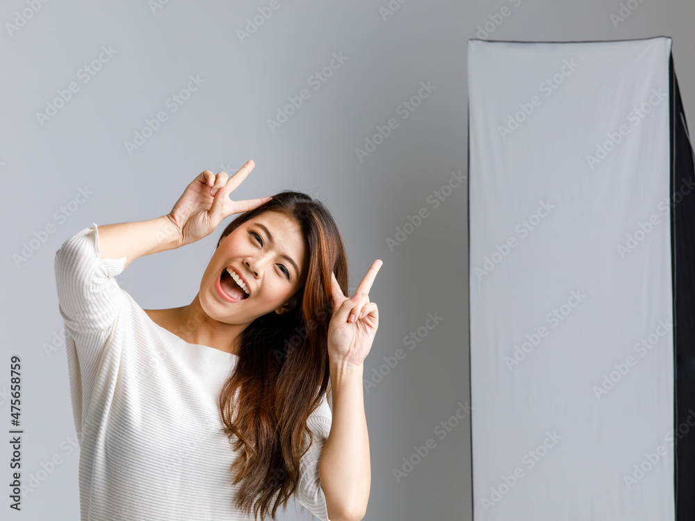 Asian young happy cheerful long hair female model in casual outfit standing smiling holding hands up showing two fingers peace sign at head in studio photography set in front lighting soft box lamp Asian young happy cheerful long hair female model in casual outfit standing smiling holding hands up showing two fingers peace sign at head in studio photography set in front lighting soft box lamp