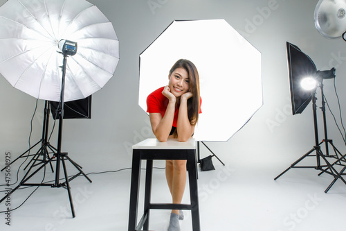 Portrait studio shot of Asian young happy cheerful female model in red casual shirt sitting on chair smiling look at camera posing crossed arms in front softbox lighting in photographing shooting set