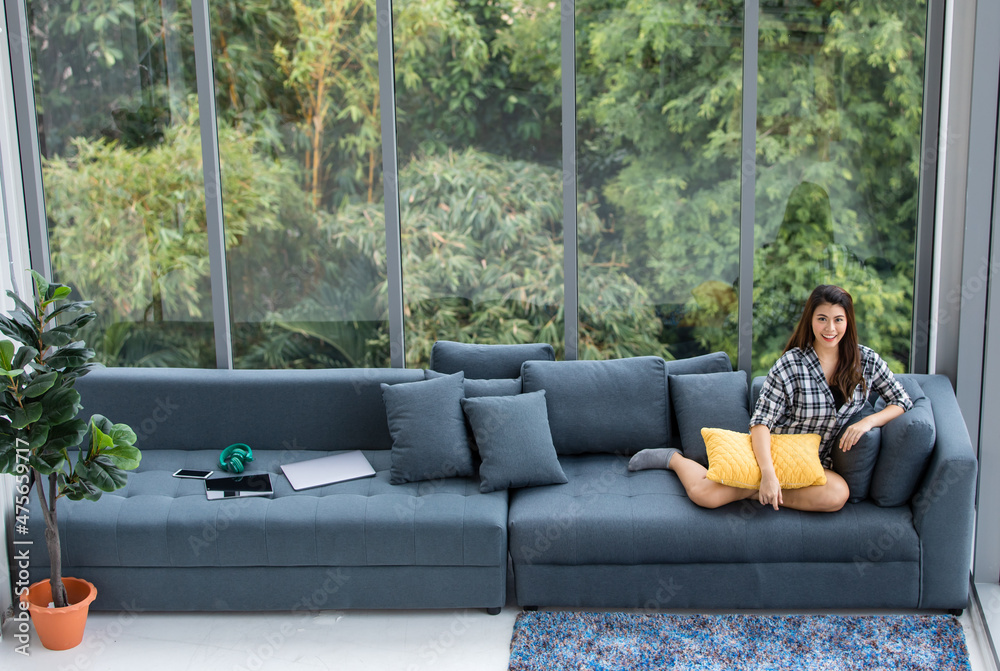 Fototapeta premium Asian pretty happy cheerful female model in plaid shirt sitting smiling on big cozy comfort sofa couch with fluffy pillows and laptop smartphone tablet headphones in front beautiful garden tree view
