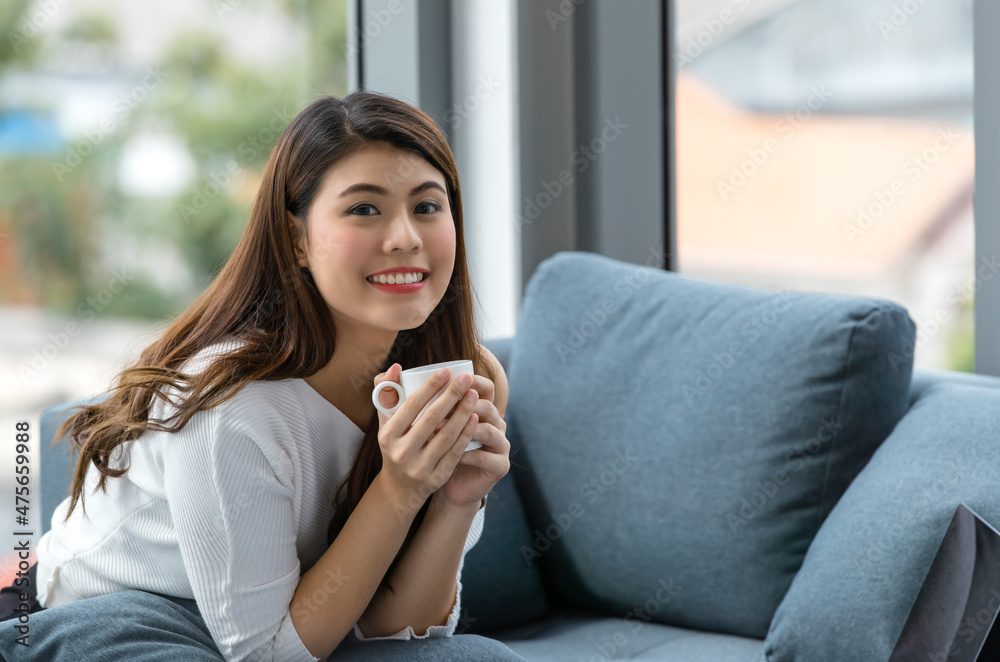 Asian happy cheerful female housewife in casual outfit sitting on cozy sofa couch with pillows resting relaxing hold drinking cup of hot coffee while browsing internet shopping online in living room Asian happy cheerful female housewife in casual outfit sitting on cozy sofa couch with pillows resting relaxing hold drinking cup of hot coffee while browsing internet shopping online in living room