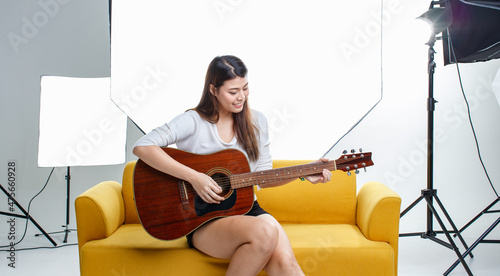 Portrait studio shot of Asian pretty female model in casual outfit sitting on cozy yellow sofa couch playing guitar on photographing shooting backstage while producer holding video movie clapperboard