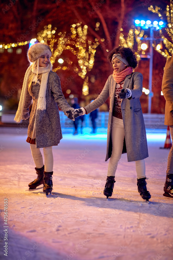 Two female friends are holding hands while skating at ice-skating rink during Christmas holidays in the city. Christmas, New Year, holiday, friendship