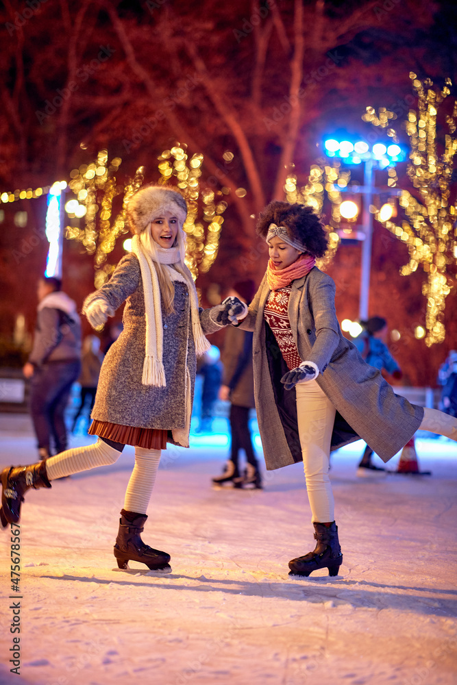 Two female friends are posing for a photo while skating at ice-skating rink during Christmas holidays in the city. Christmas, New Year, holiday, friendship