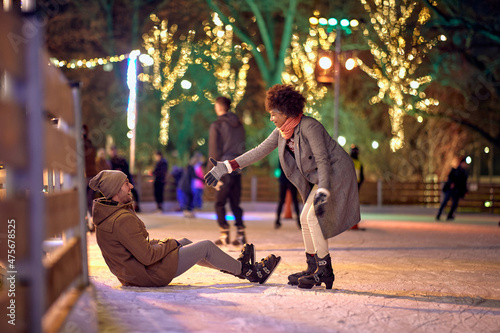 A girl is helping her boyfriend to stand up while learning to skate at ice-skating rink during christmas holidays in the city. Christmas, New Year, holiday, love