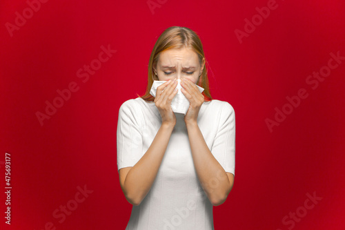 Unhappy sick young girl suffer cold or allergy, feeling unwell, wiping runny nose by napkin on red studio background
