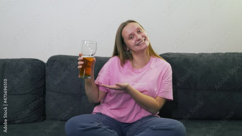Young caucasian woman in pink T-shirt is sitting on gray sofa at home and drinking beer from transparent glass. She laughs and smiles. Beer advertising. Bad habit, alcohol, alcoholism, unhealthy diet Young caucasian woman in pink T-shirt is sitting on gray sofa at home and drinking beer from transparent glass. She laughs and smiles. Beer advertising. Bad habit, alcohol, alcoholism, unhealthy diet