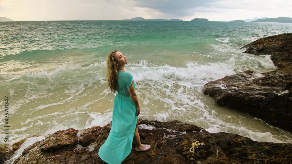 Woman walks on rock of sea reef stone, stormy cloudy ocean. Blue swimsuit dress tunic. Concept rest, tropical resort coastline tourism summer holidays Woman walks on rock of sea reef stone, stormy cloudy ocean. Blue swimsuit dress tunic. Concept rest, tropical resort coastline tourism summer holidays