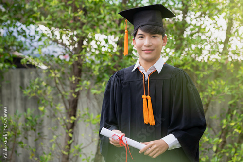 In the graden, a happy young Asian man wears graduation gowns and has a diploma.