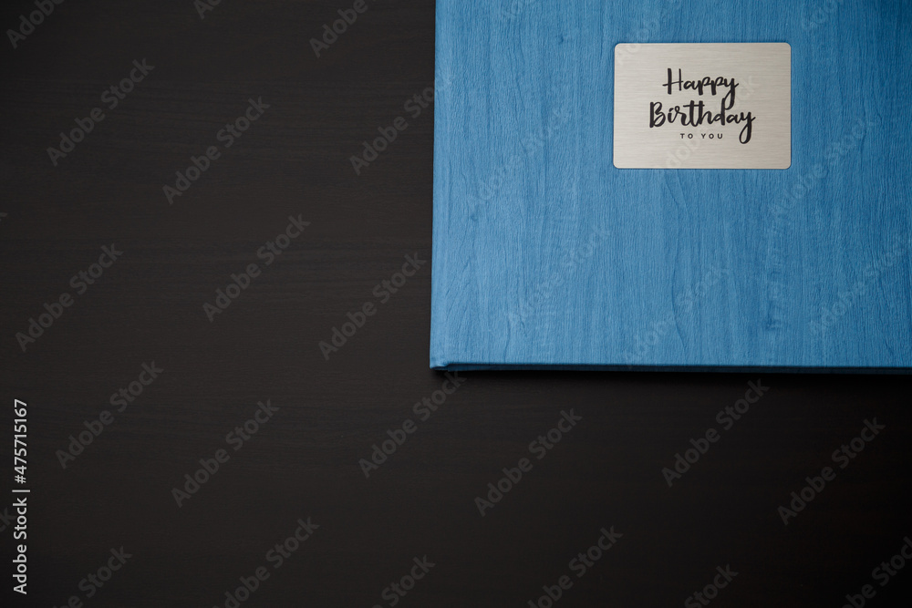 Obraz premium Photobook, photoalbum in blue leather cover on black table background with metallic shield and inscription Happy Birthday to you. Top view, copy space.