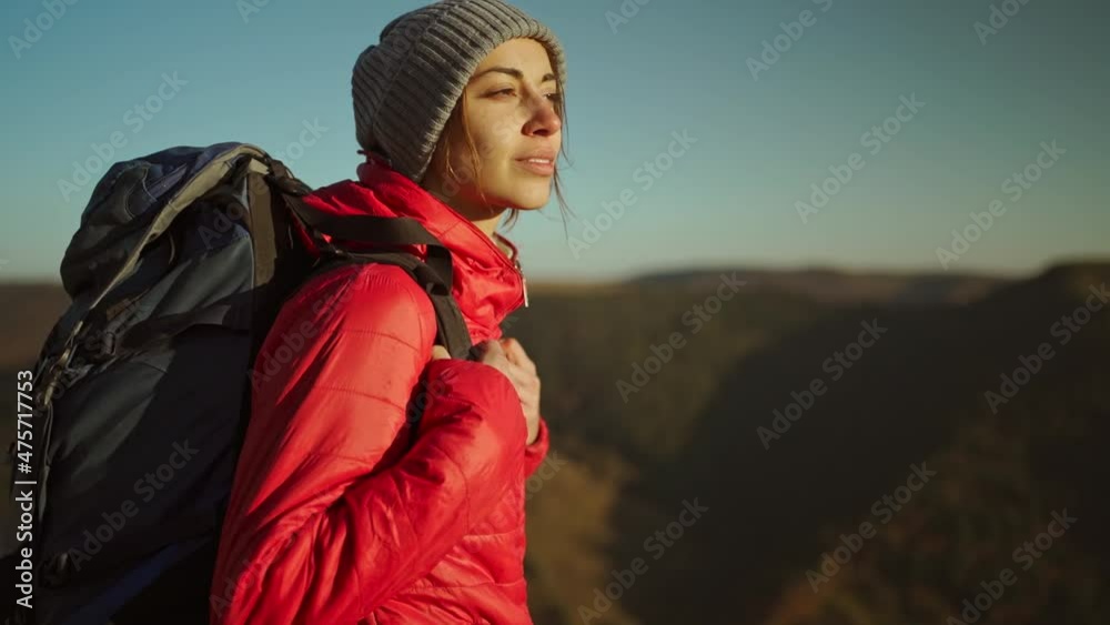 slow motion portrait woman hiker in bright red jacket with backpack stands on mountain top against background of sunset sky over mountains, looking ahead and enjoying freedom and successful ascent.