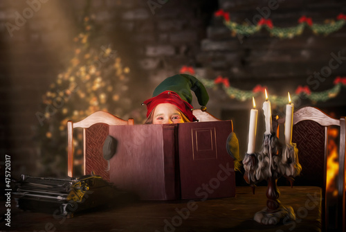 a girl with bright beautiful eyes sits at home at a table for the new year and Christmas stroking out from behind a book in the costume of the elf assistant santa claus for 2022