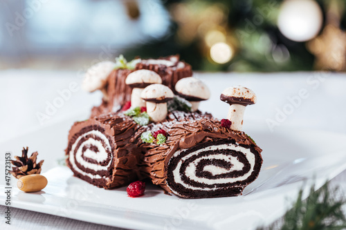 French dessert called Yule log or bûche de Noël with merengue mushrooms and mint leaves on top of chocolate glazing. Placed in front of Christmas tree. Decorated for Christmas Holidays or New year