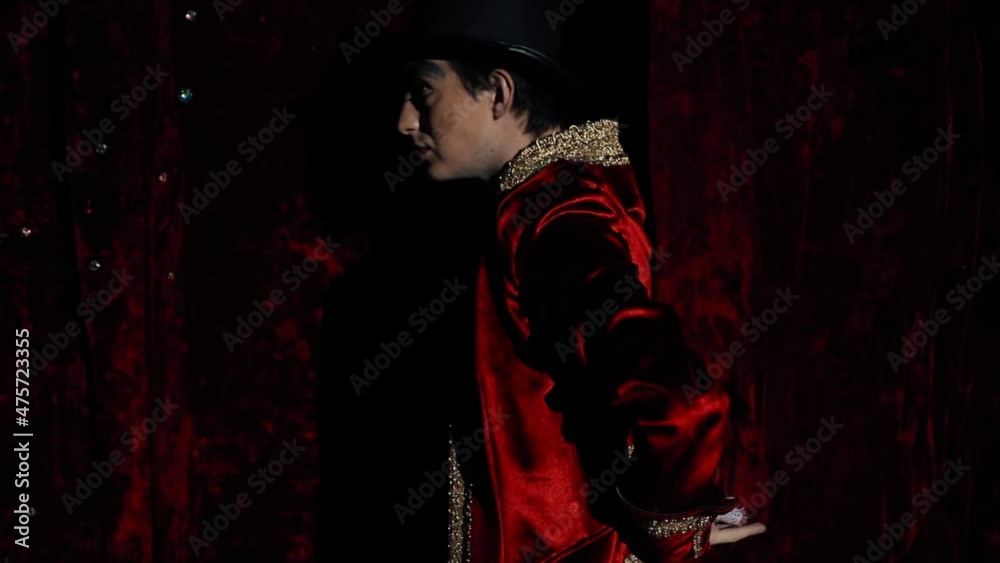 The showman effectively begins to show the trick with his fingers, passes through the curtain, enters the stage. The guy is a wizard with makeup in a red dress coat and top hat. A bright suit against The showman effectively begins to show the trick with his fingers, passes through the curtain, enters the stage. The guy is a wizard with makeup in a red dress coat and top hat. A bright suit against