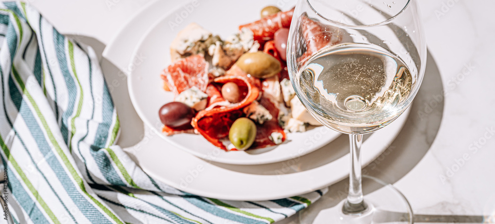 cool white wine in a glass near a plate with sliced jamon, dove cheese and various olives. a light snack on a hot day. classic italian food.