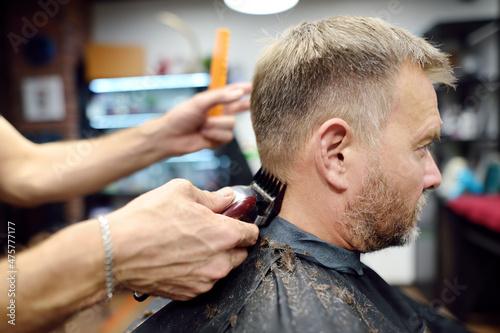 Hairdresser is cutting hair of handsome bearded mature man in salon. Stylist making hairstyle with electric shaver for person in barbershop. Services of a professional stylist.
