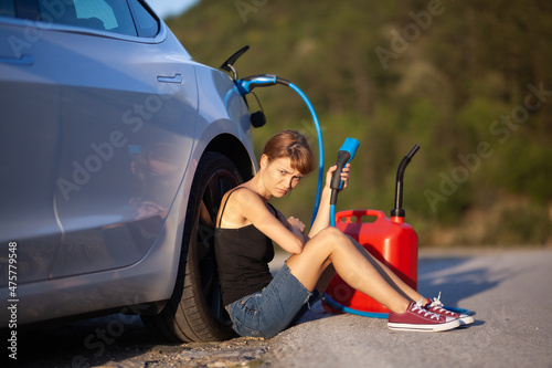 Sad girl sitting on the ground next to electric car. Holding charging cable and red gassoline canister photo