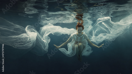 Fotografering A girl in a white dress posing underwater on a dark background as if she were in