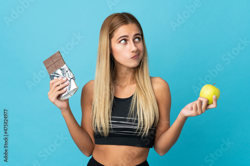 Young blonde woman isolated on blue background having doubts while taking a chocolate tablet in one hand and an apple in the other