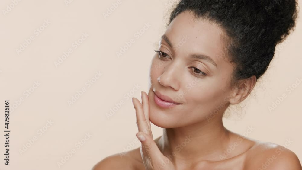 Young slim pretty black-haired African American woman with naked shoulders strokes her face and turns her head to the camera smiling wide for it on beige background | Smooth face concept