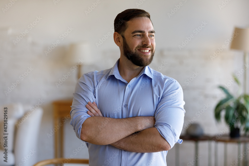 Obraz premium Happy confident young businessman entrepreneur manager standing with folded arms at home office, looking in distance visualizing future, imagining opportunities or thinking of company development.