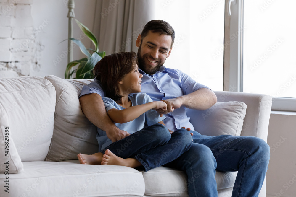 Smiling preteen small kid boy bumping fists with joyful caring young father, making decision, enjoying talking speaking communicating, resting on comfortable sofa together on weekend at home. Smiling preteen small kid boy bumping fists with joyful caring young father, making decision, enjoying talking speaking communicating, resting on comfortable sofa together on weekend at home.