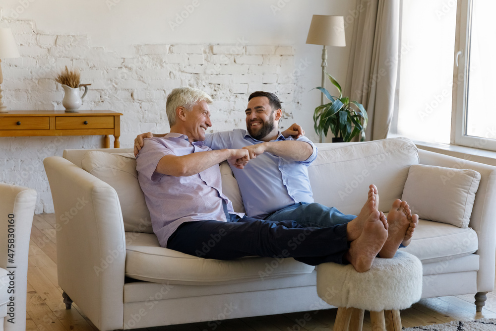 Happy bonding mature man bumping fists with grownup son, relaxing together on sofa with feet on footstool, having fun enjoying trustful conversation, different generations family relations concept. Happy bonding mature man bumping fists with grownup son, relaxing together on sofa with feet on footstool, having fun enjoying trustful conversation, different generations family relations concept.