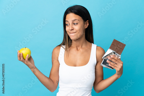 Caucasian girl isolated on blue background having doubts while taking a chocolate tablet in one hand and an apple in the other