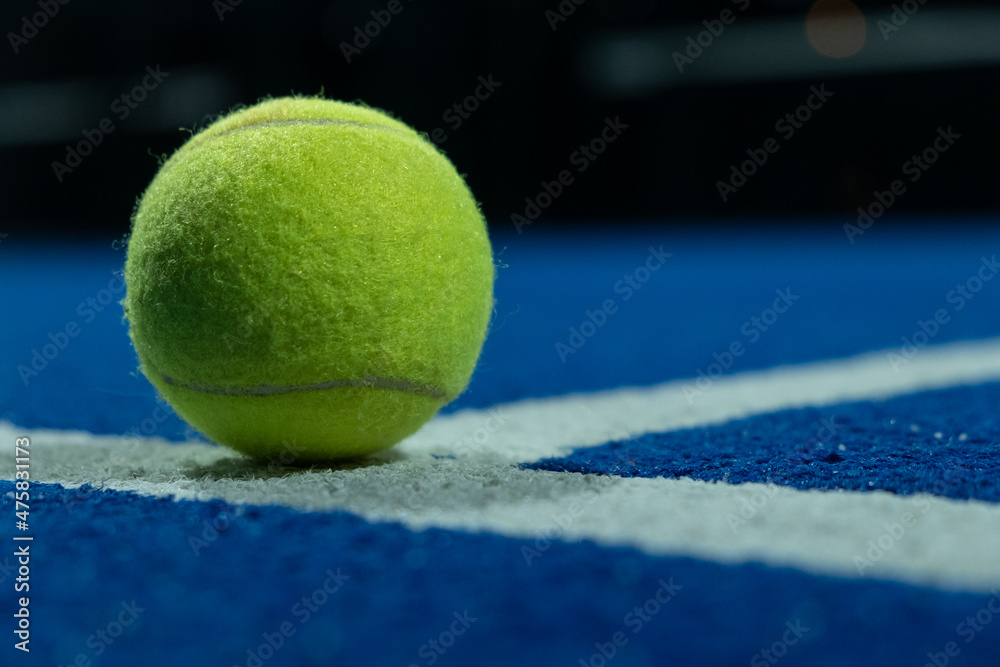 A paddle tennis ball in the foreground on the line of a blue paddle tennis court at night.