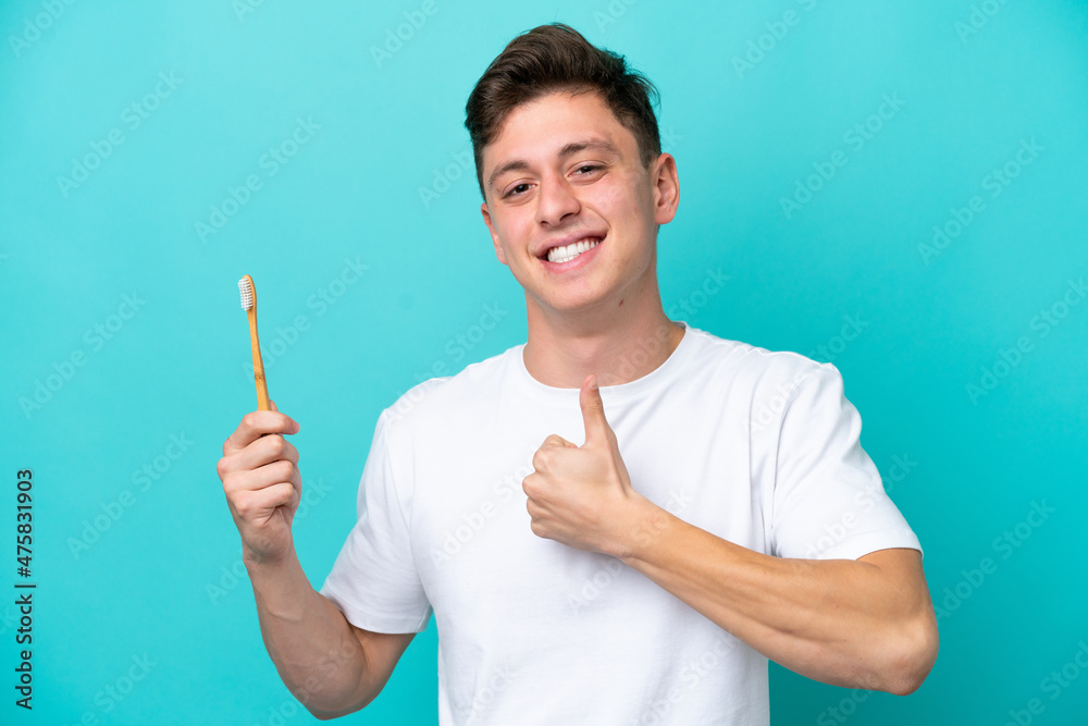Young Brazilian man brushing teeth isolated on blue background giving a thumbs up gesture Young Brazilian man brushing teeth isolated on blue background giving a thumbs up gesture