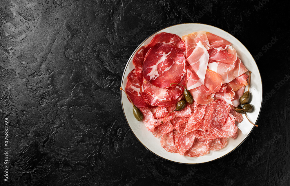 Traditional spanish jamon, prosciutto crudo, italian salami, parma ham. Antipasto plate and capers. Black  background. Top view.