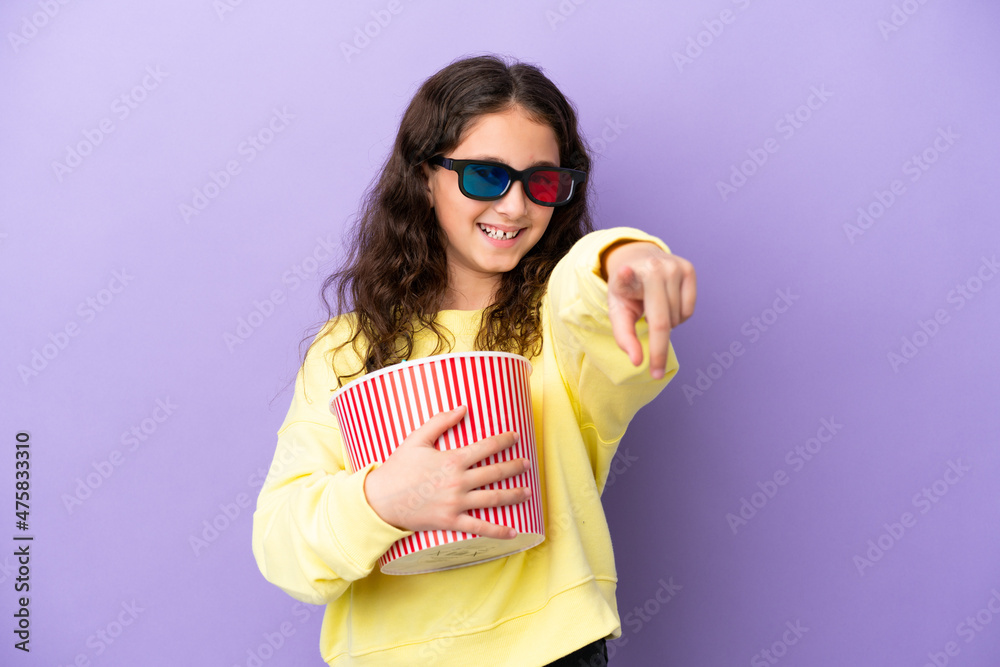 Little caucasian girl isolated on purple background with 3d glasses and holding a big bucket of popcorns while pointing away