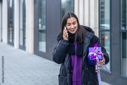 A young woman with a purple gift in her hands talking on the phone, outside. © puhimec
