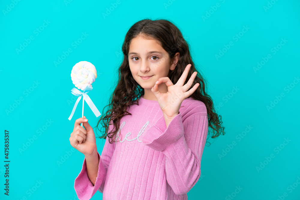 Little caucasian girl holding a lollipop isolated on blue background showing ok sign with fingers Little caucasian girl holding a lollipop isolated on blue background showing ok sign with fingers