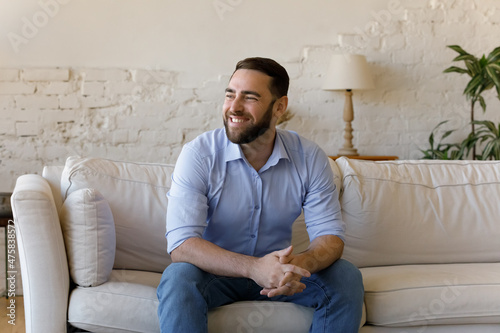 Obraz na plátně Happy dreamy millennial generation male homeowner looking in distance sitting on cozy couch, visualizing future or feeling excited thinking of good news, relaxing alone in modern living room