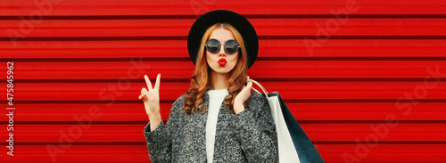 Fashionable portrait of stylish young woman model posing with shopping bags and blowing her lips sending sweet air kiss wearing gray coat, hat on colorful red background © rohappy