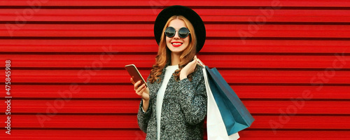 Fashionable portrait of happy smiling young woman with smartphone and shopping bags wearing a gray coat, hat on colorful red background © rohappy