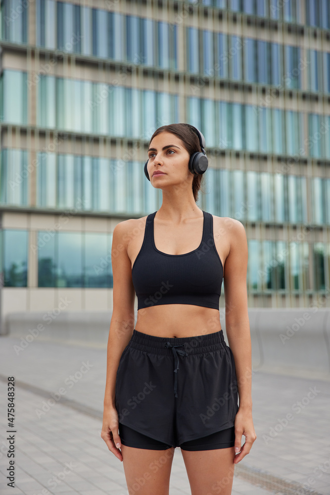 Sporty woman has regular training outdoors poses in urban setting wears black tracksuit listens music via headphones concentrated into distance thoughtfully leads healthy lifestyle. Fitness model