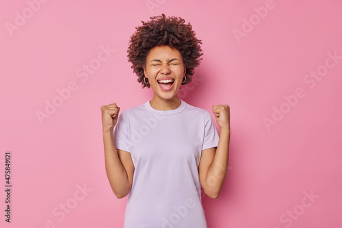 Waist up shot of overjoyed curly woman clenches fists celebrates success feels like winner exclaims happily wears casual t shirt isolated over pink background finally catches dream. Yes gesture