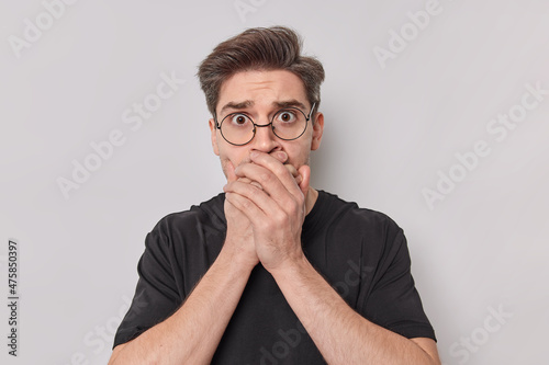 Shocked speechless young European man covers mouth with hands ties to be mute looks with fear at camera wears spectacles and casual t shirt poses against white background. Keep silence concept