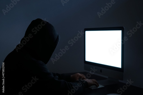 unidentified hacker wearing a black hoodie is sitting in front of the white blank screen. a man initiating a cyber attack confidentially. photo