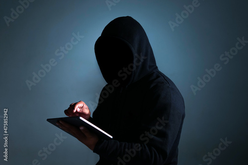 a mysterious man wearing a black hoodie is holding and operating a tablet. a portrait of an anonymous hacker doing a cybercrime. photo