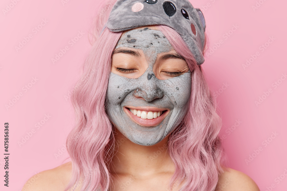 Headshot of happy gentle woman with pink hair keeps eyes closed applies clay mask undergoes beauty treatments wears sleepmask stands topless poses indoor. Skin care and cosmetic procedures concept